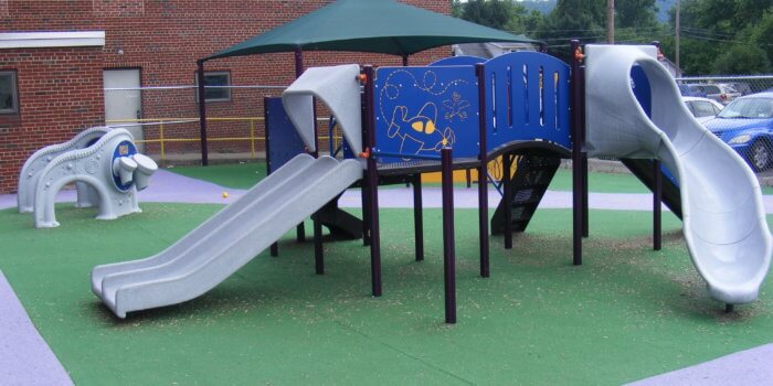 Photo of a play structure with slides, panels, and climbers, with sensory panels and a shade structure in view behind