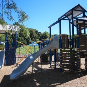 Photo of two play structures, each with a roof, slides, and climbers, and connected with a net climbing bridge