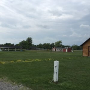 Photo of the site before playground installation; an empty field with some sheds and play equipment visible farther on