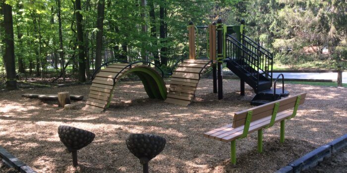 Photo of nature inspired playground, with a faux wood bridge, acorn seats, and a log balance beam.