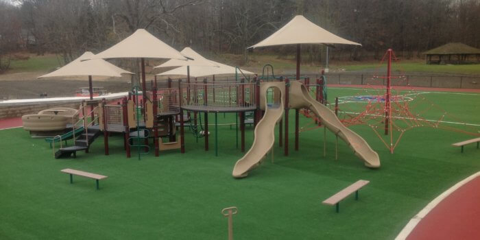 Photo of sprawling play structure with multiple decks, slides, and a wheelchair accessible glider, as well as shades and a rope climbing structure.