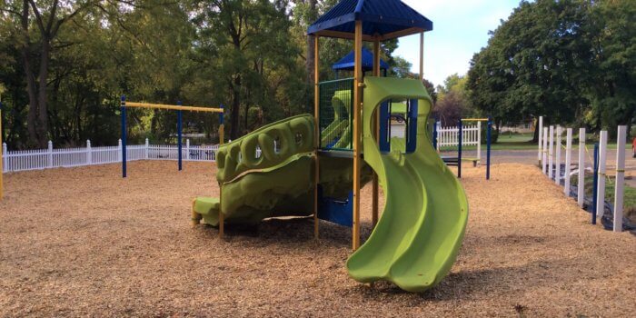 Photo of two play structures, each with climbers, a slide, and a roof, as well as a bay of swings and an independent spring rider.