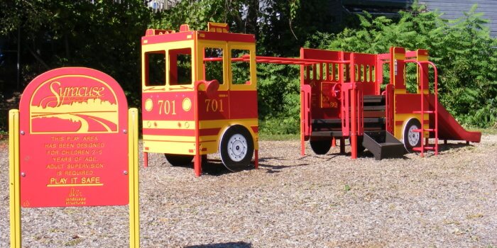 Photo of small custom firetruck-shaped playground, with climbers, play panels, and a slide.
