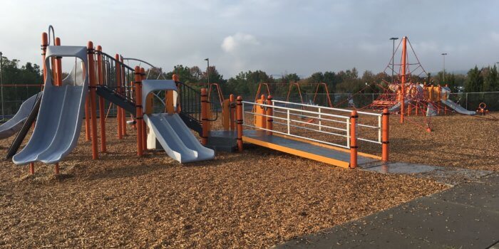 Photo of play structure with multiple slides, climbers, and a wheelchair ramp, and a rope climbing structure in the background.